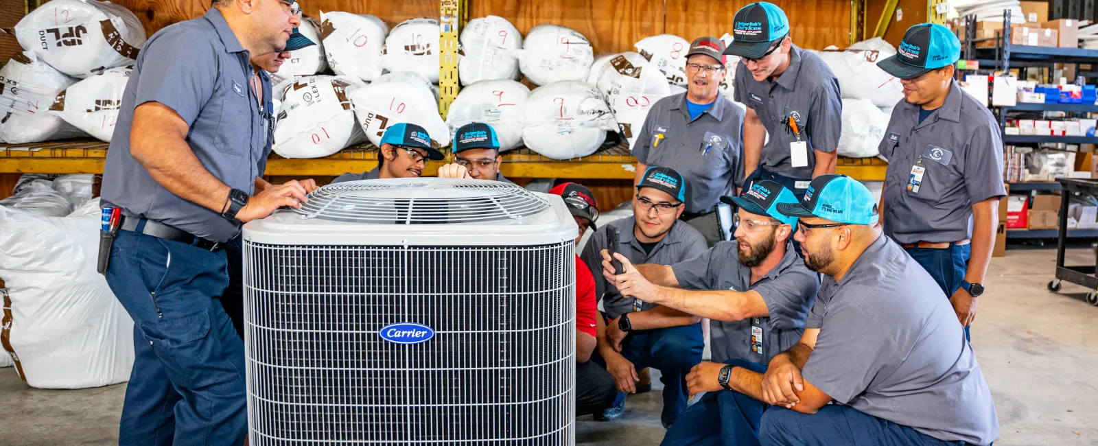 Group of technicians in uniform inspecting a Carrier HVAC unit in a warehouse filled with supplies and air filters.