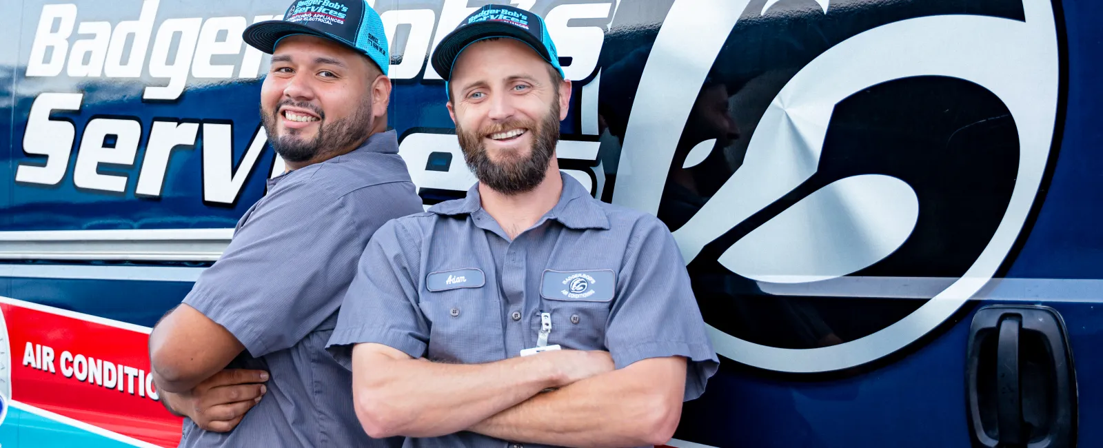 Two smiling HVAC technicians in uniform standing back-to-back by a Badger Heating Services van.