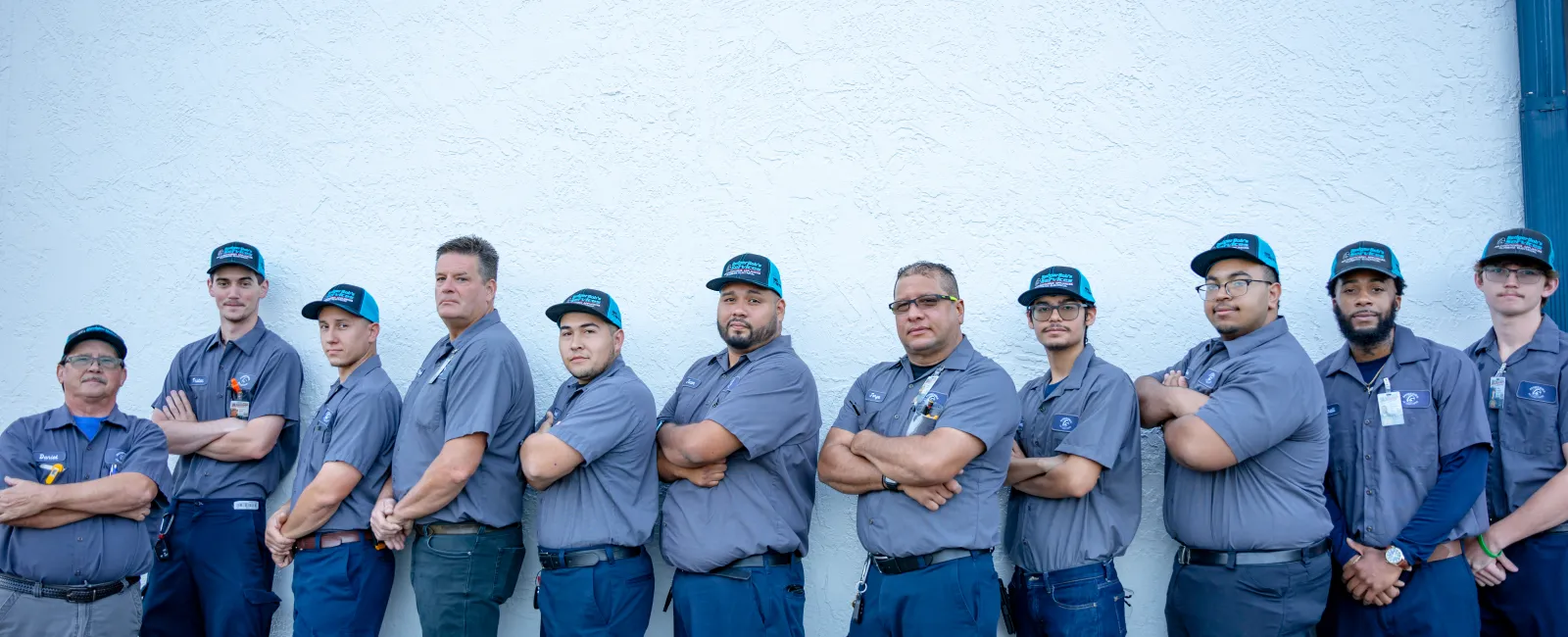 Group of eleven uniformed male workers standing side by side against a white wall with arms crossed.