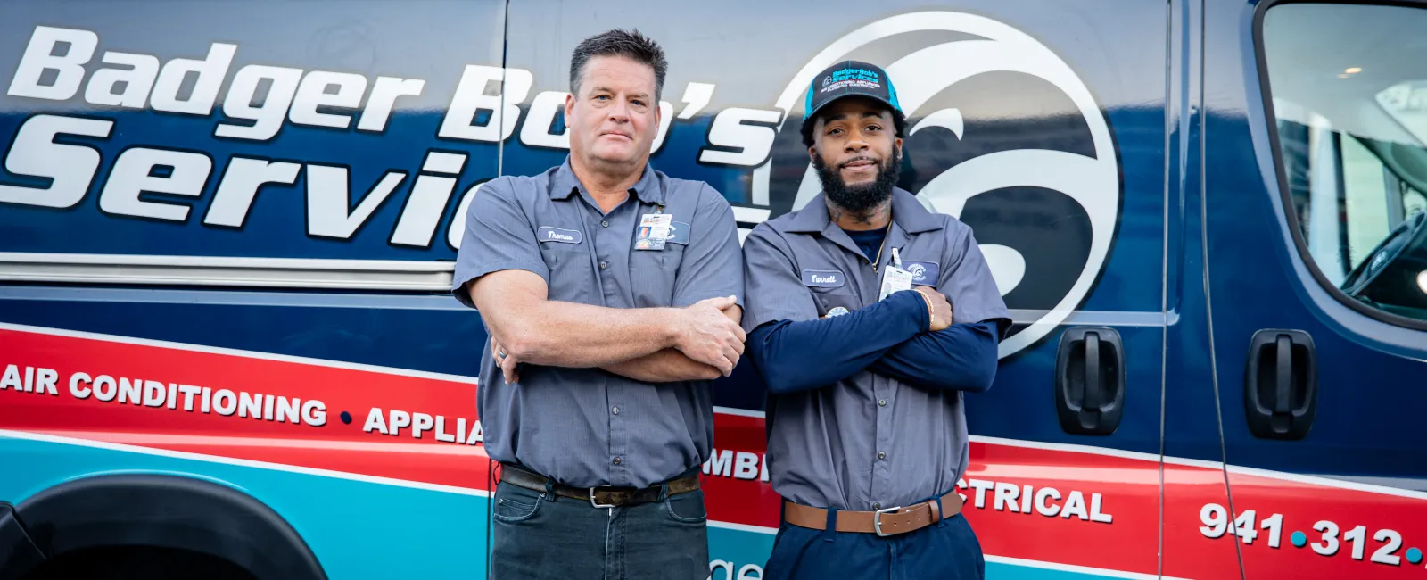Two uniformed technicians standing with arms crossed in front of a Badger Boy's Services company van with equipment on top.