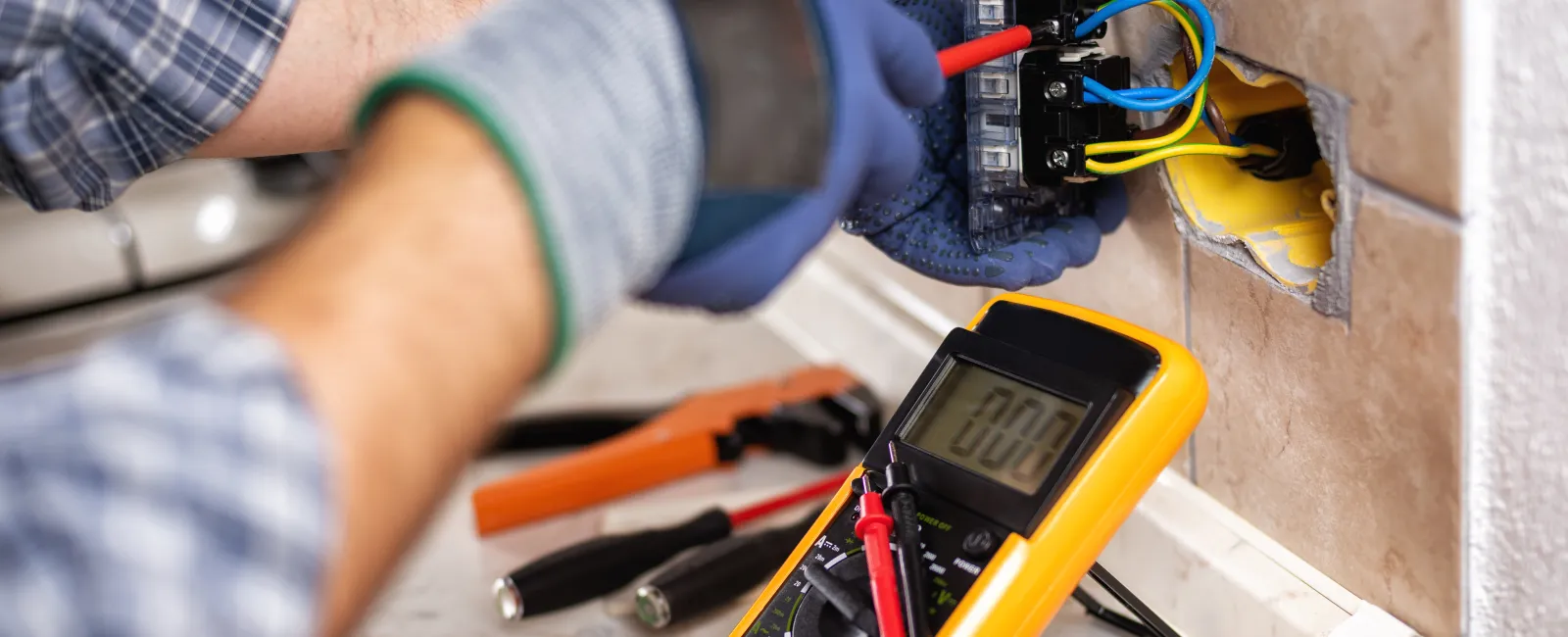 Electrician wearing gloves working on wall wiring using a multimeter and tools on a marble surface.
