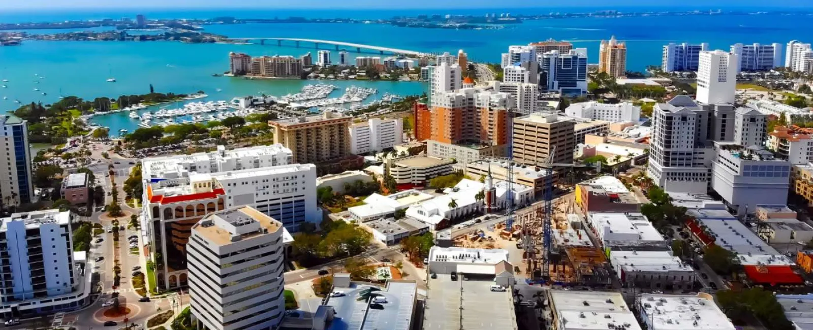 Aerial view of a coastal city with high-rise buildings, marina, bridge, and blue ocean under clear sky.