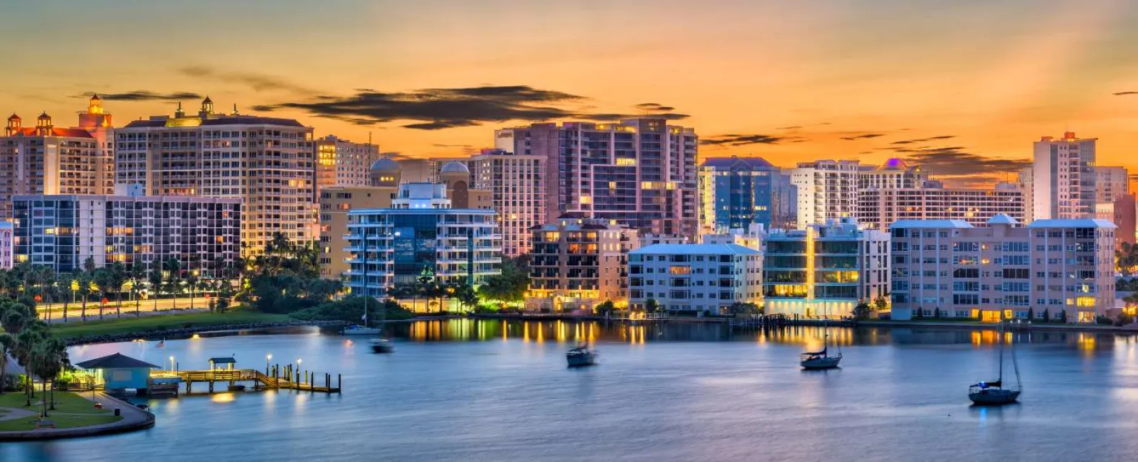 City skyline at sunset with illuminated buildings and boats on calm water near the shore