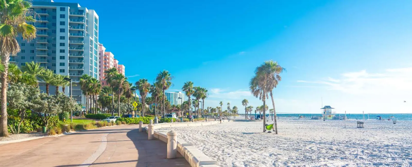Sunny beach with white sand, palm trees, skyscrapers, boardwalk, and clear blue sky by the ocean.