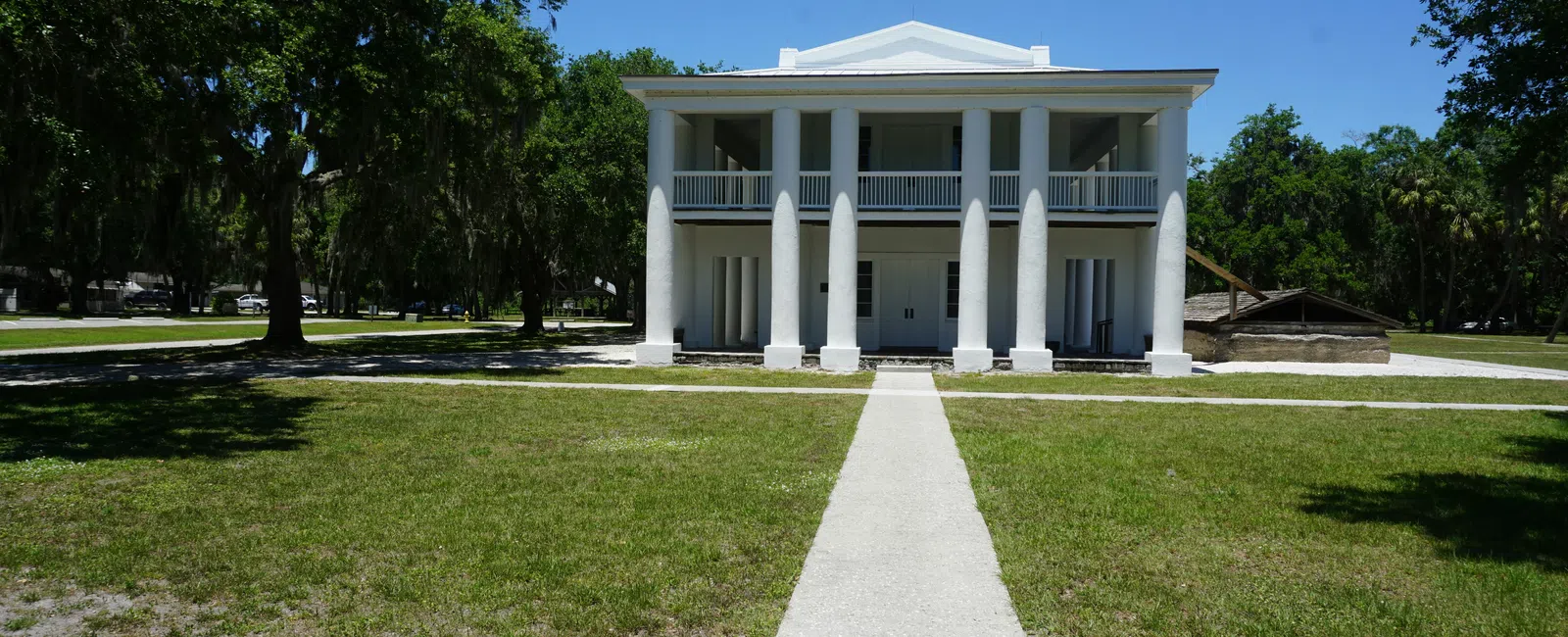 White historic Ellenton Gamble Plantation two-story building with columns, surrounded by green lawn and trees under clear blue sky representing we have HVAC near you