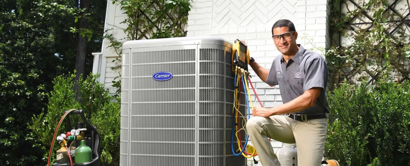 HVAC technician servicing Carrier air conditioning unit outside a home in a green yard with tools and safety glasses.