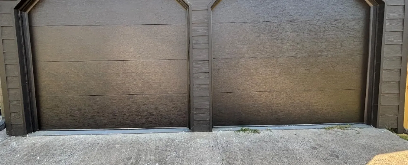 Two closed brown garage doors with a concrete driveway and exterior wood siding around.