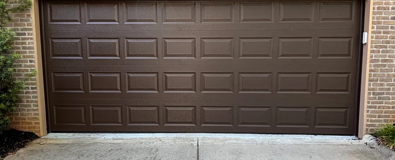 Closed brown paneled garage door set in brick house with concrete driveway and pitched roof.