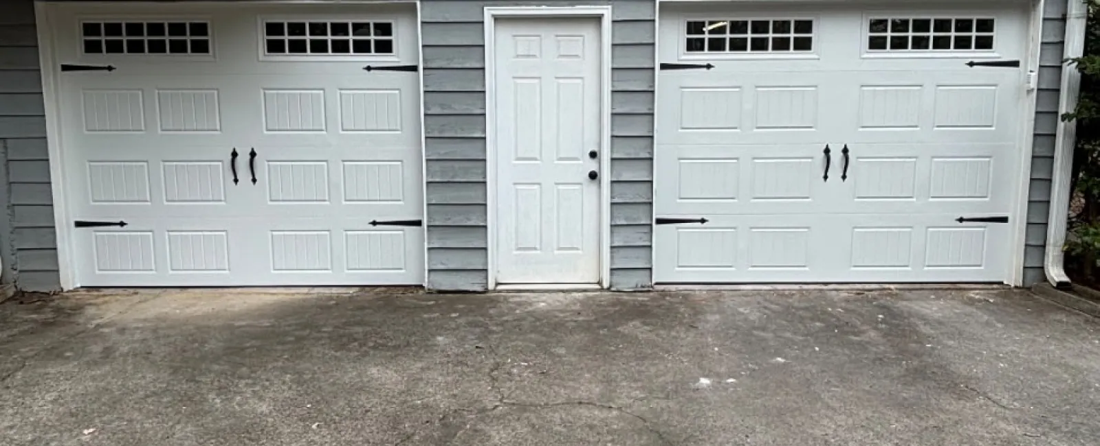 Two white garage doors with black hardware and a white door in the middle on a gray house exterior.