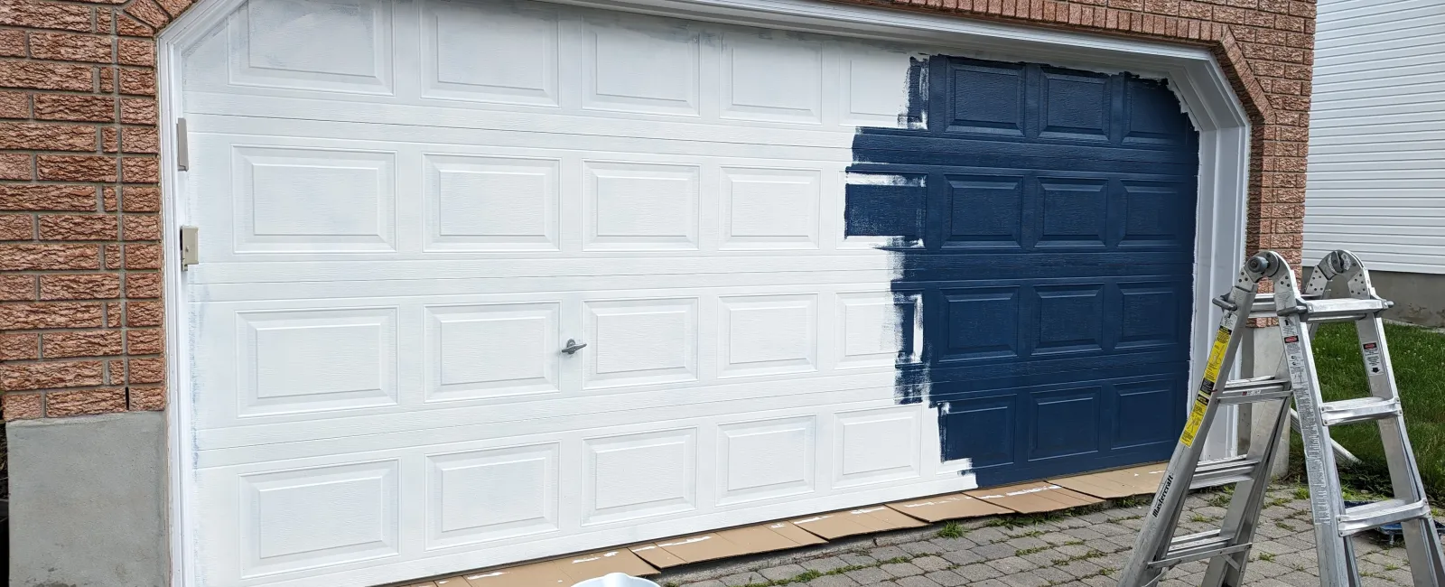 Partially painted garage door with dark blue paint, white base coat, ladder, bucket, and roller on driveway.