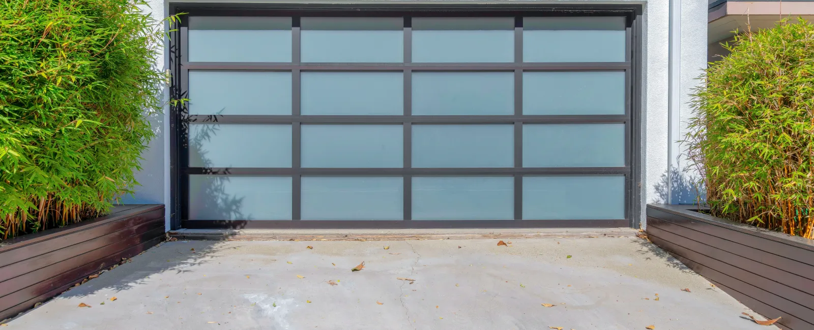 Modern frosted glass garage door with black frame in a white house exterior and greenery on both sides
