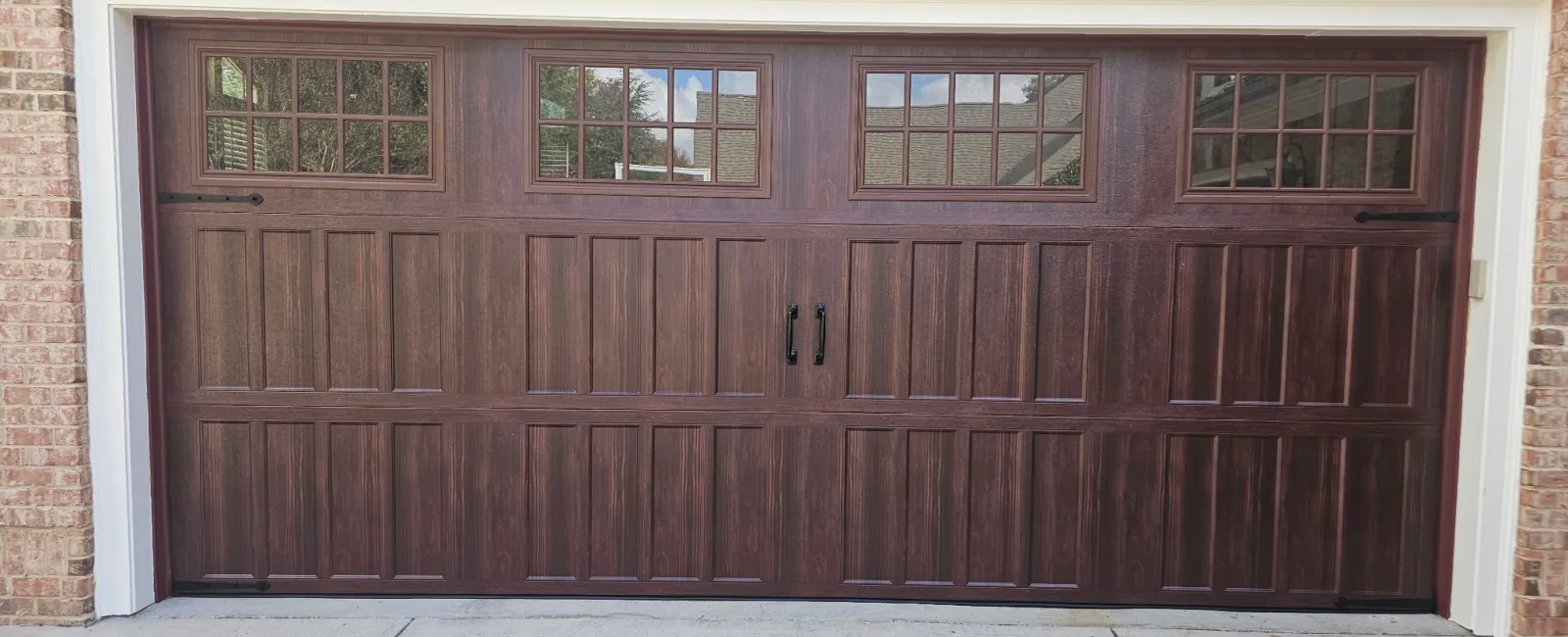 Dark wood garage door with window panels and black handles set in a brick house exterior with concrete driveway.