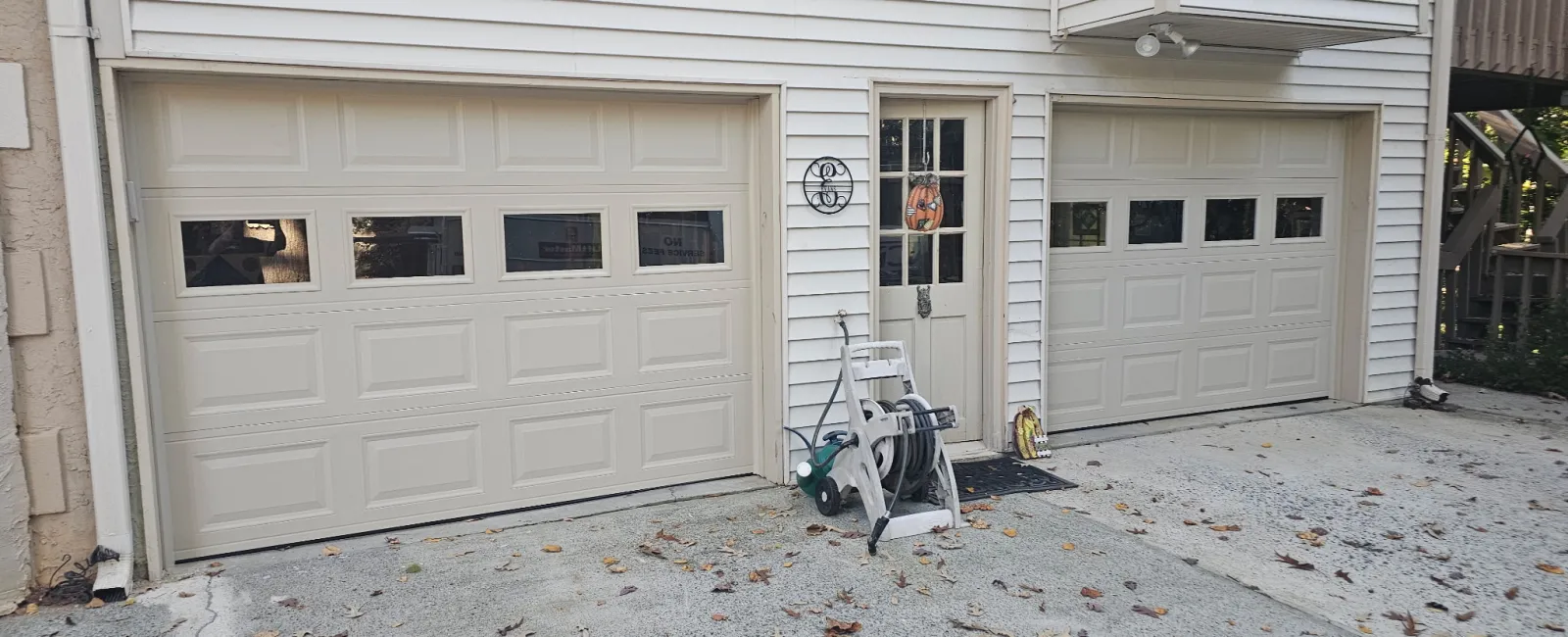 Two beige garage doors with windows and a central door on a white house exterior with a hose reel nearby.