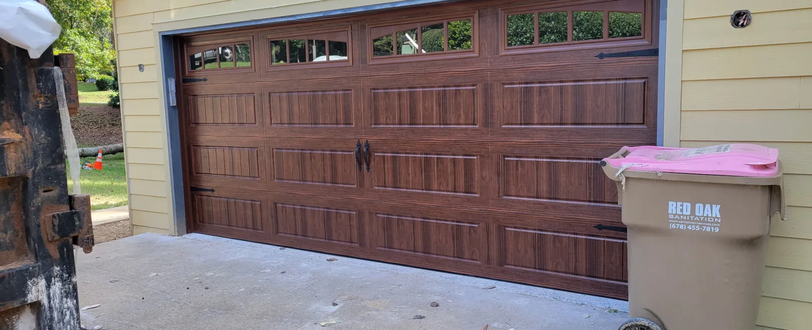 New wooden garage door with windows installed on beige house exterior with driveway and trash bin.