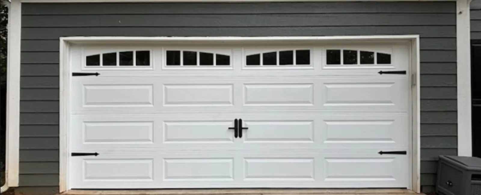 White paneled garage door with small upper windows, black decorative handles, and gray siding exterior.