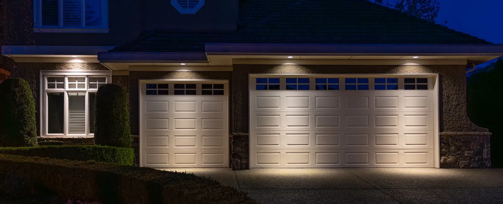Suburban house with two illuminated garage doors at night and well-manicured bushes in driveway area.