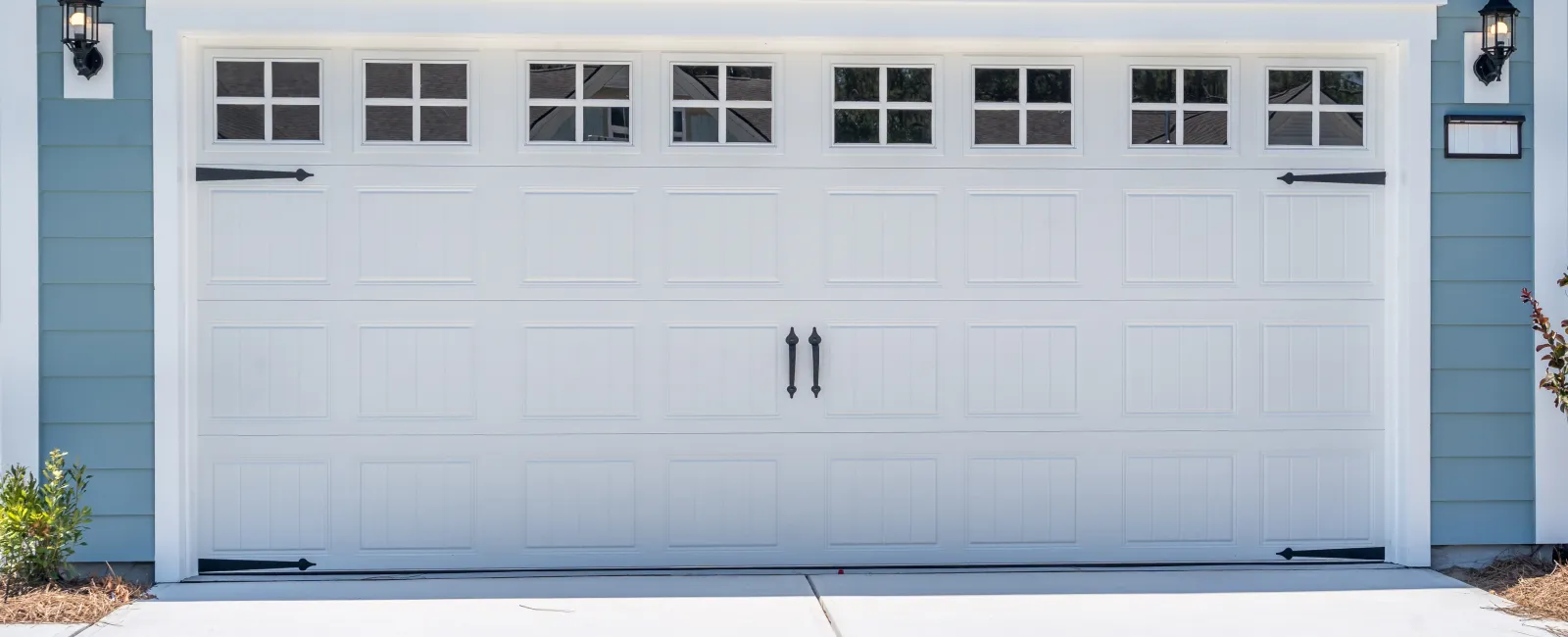 White paneled garage door with windows and black handles on a blue house exterior with concrete driveway.