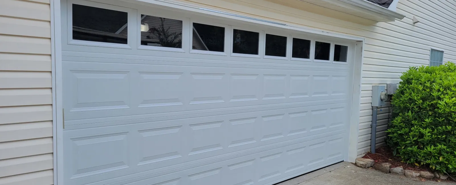 White residential garage door with windows at the top and beige siding on a suburban home.