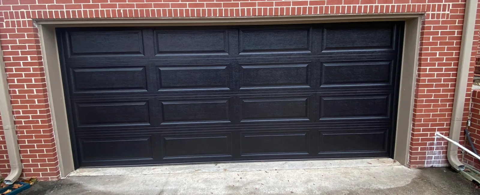 Black paneled garage door set in a red brick house facade with two windows and a concrete driveway.