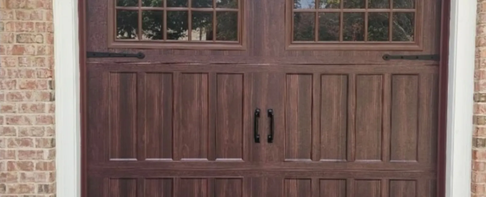 Dark wood paneled garage door with windows and decorative black handles on brick house exterior