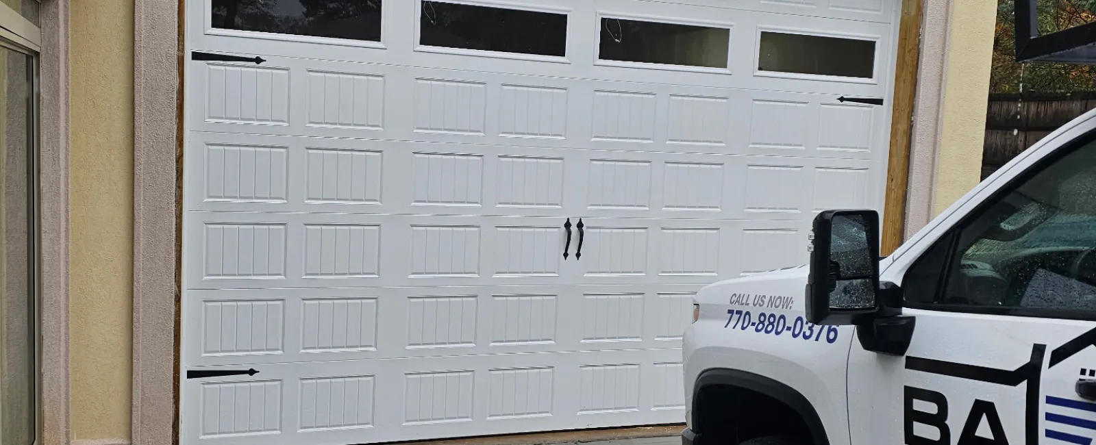 Freshly installed white garage door on beige house with service truck parked outside on wet driveway.