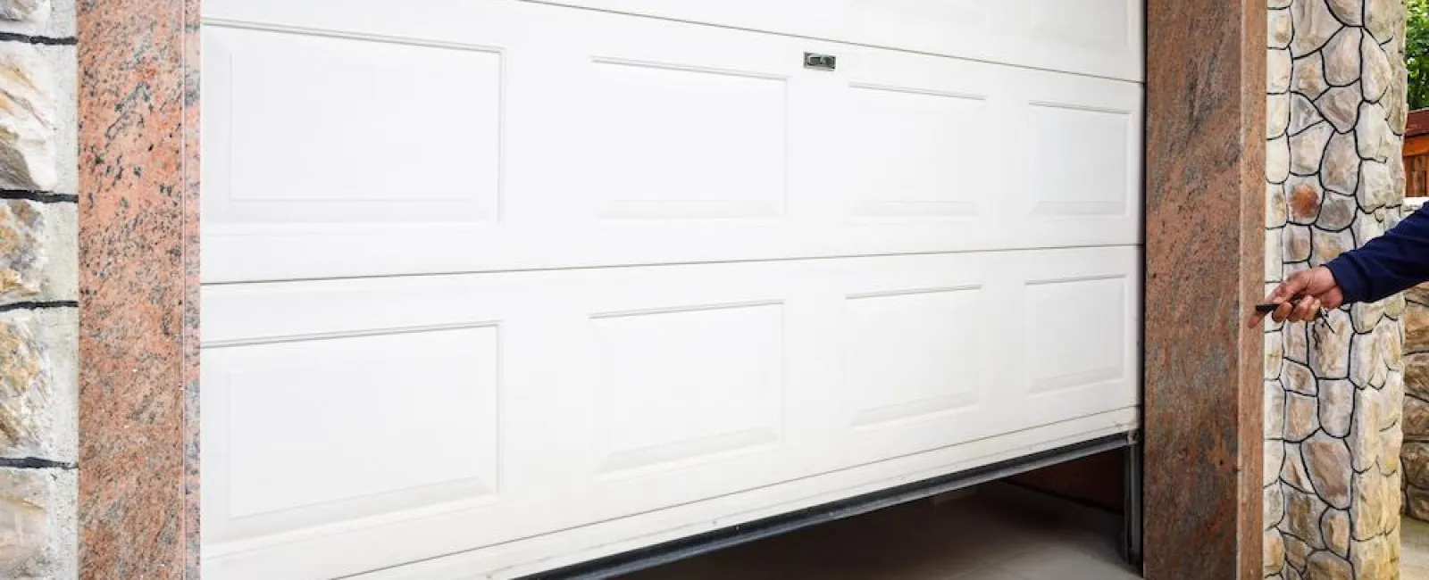 Person opening a white panel garage door in a stone-walled driveway entrance.