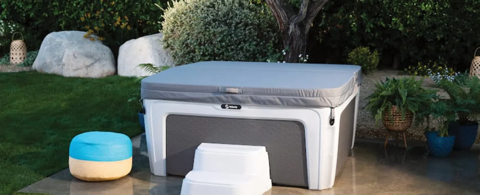 Modern hot tub with gray cover and white steps beside a blue and beige pouf in a green garden patio.
