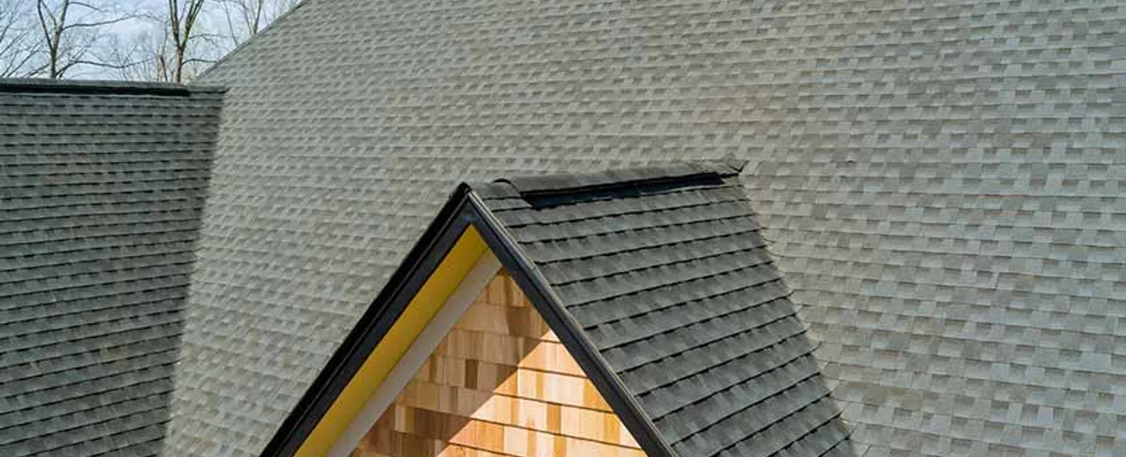 Close-up of gray shingled roof with a dormer window featuring wooden siding under clear sky