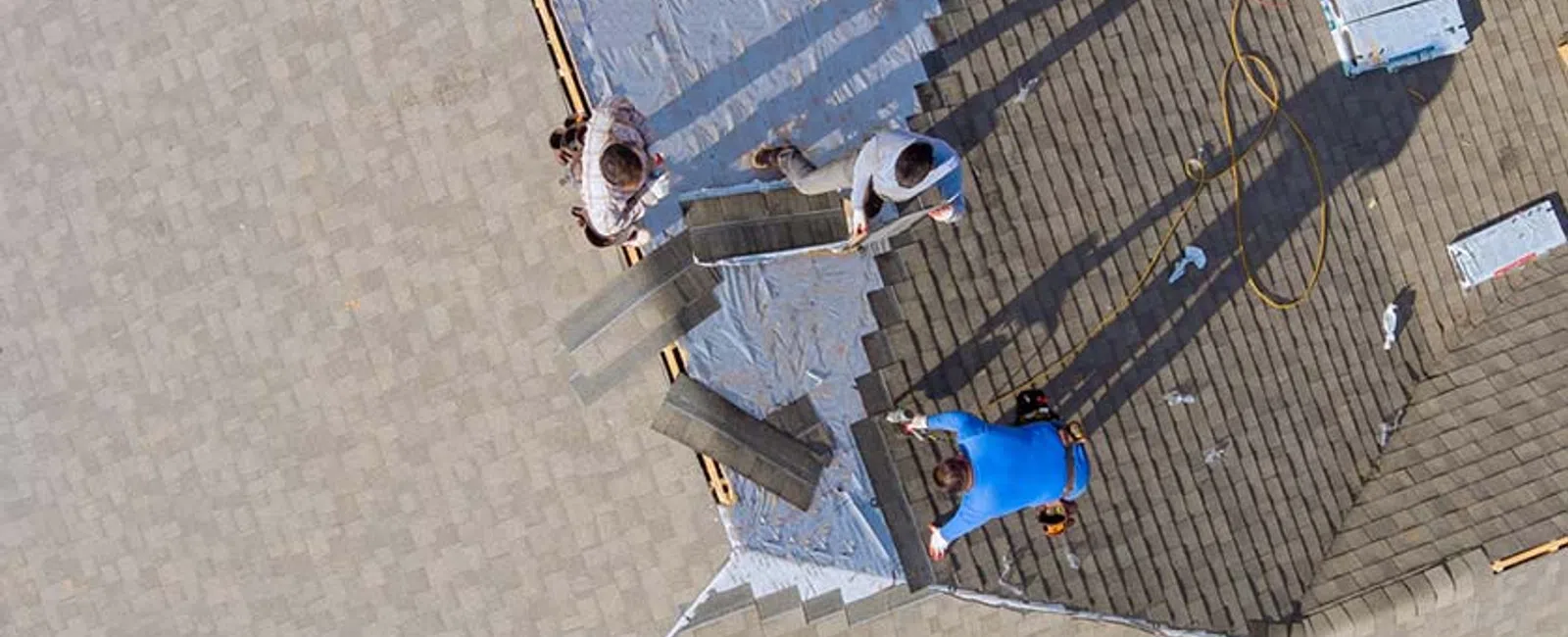 Aerial view of three workers installing roofing shingles on a house roof under sunny conditions.
