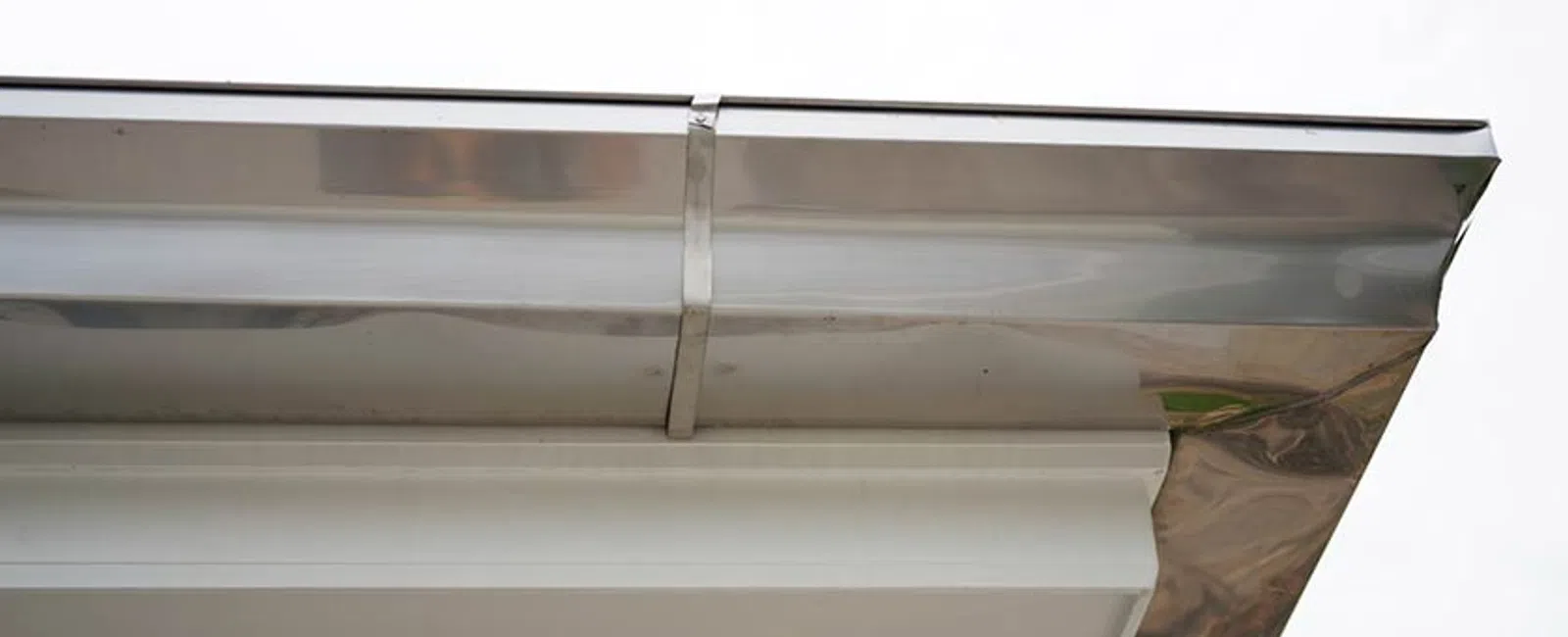Close-up of a modern stainless steel rain gutter installed on a white building roof edge under cloudy sky.