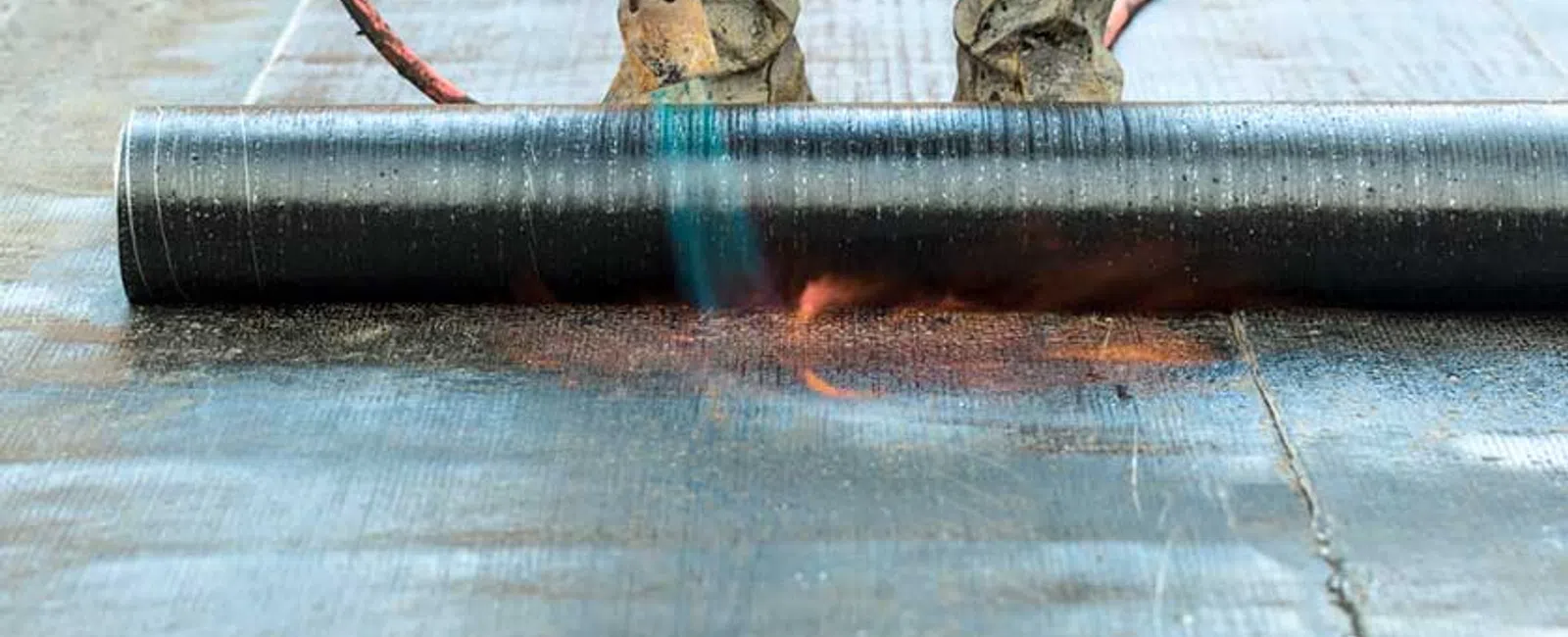 Worker using a blowtorch to apply waterproofing membrane on a flat roof surface wearing protective boots.