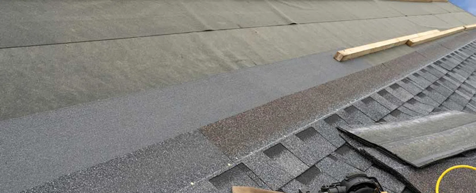 Roof under construction with asphalt shingles, wooden battens, nail gun, and tool pouch on a clear day.