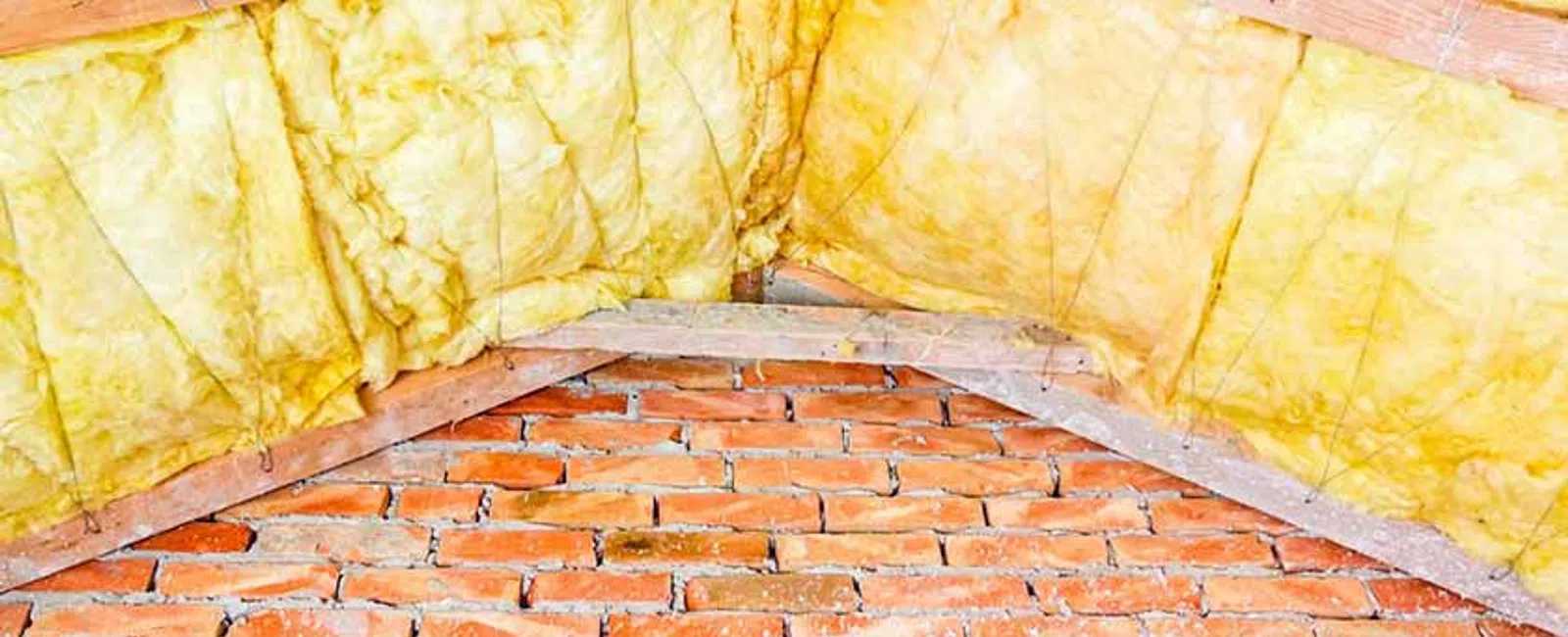 Yellow fiberglass insulation installed between wooden beams above a brick wall in an attic space