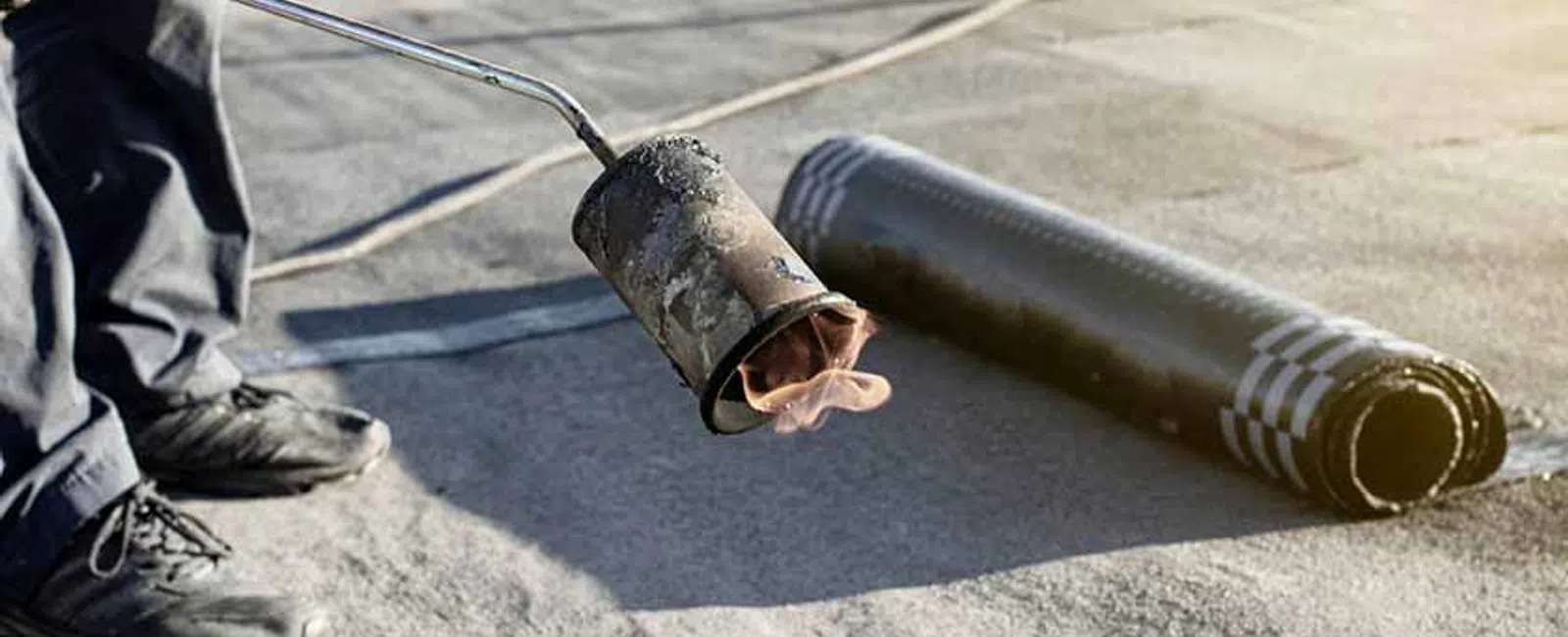 Worker using a torch to apply roll roofing material on a flat roof during installation process.