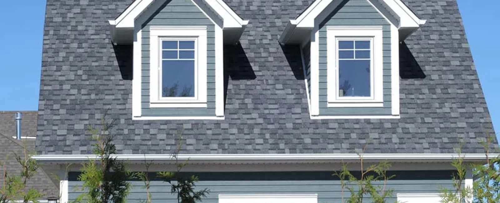 Modern blue house with gray shingled roof and white framed windows behind green shrubs under clear sky