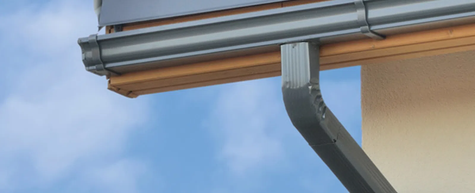 Close-up of a house roof corner with metal gutter and downspout under a clear blue sky.