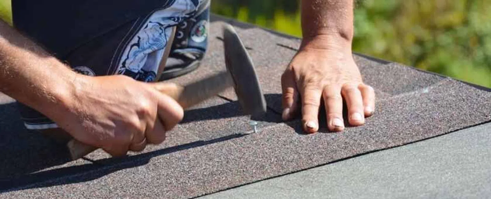 Close-up of hands hammering nails into asphalt shingles on a roof under sunny daylight.