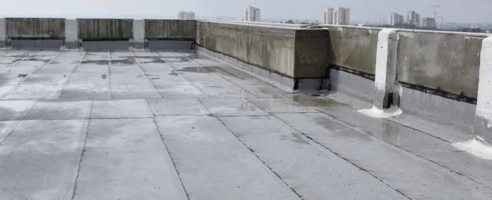 Flat rooftop with concrete barriers and waterproof roofing membrane on a cloudy day in an urban area.