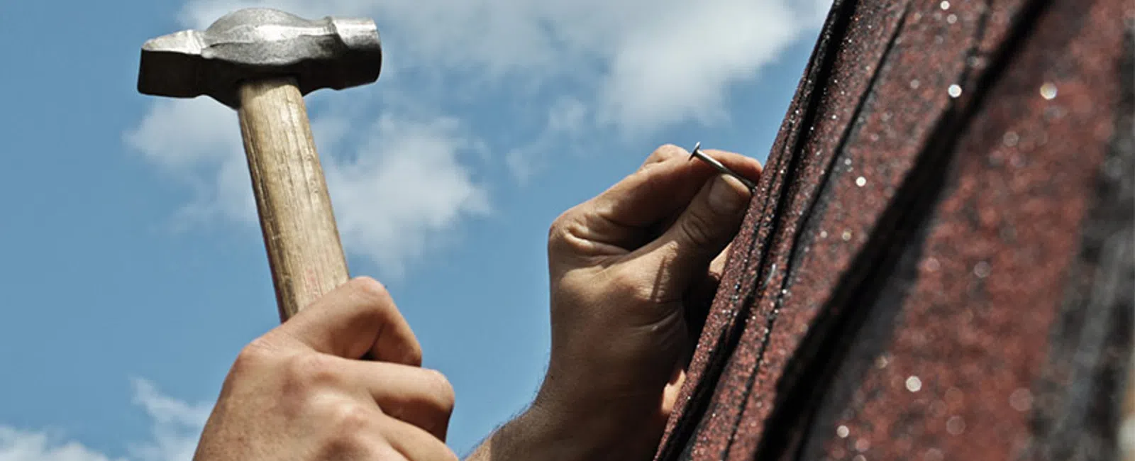 Hands using hammer to nail on textured roofing shingles under a partly cloudy sky outdoors