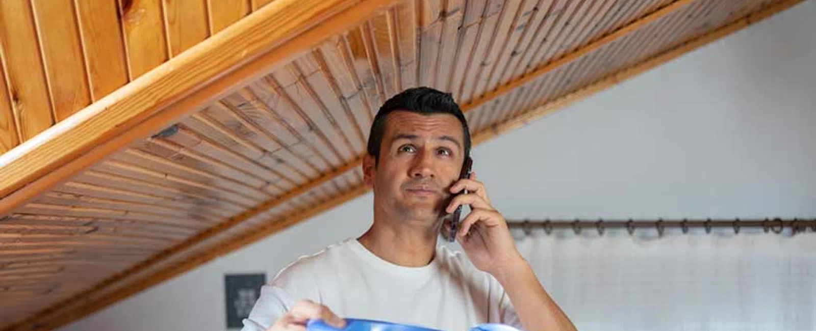 Man holding blue bucket under leaking wooden ceiling while talking on phone in living room.