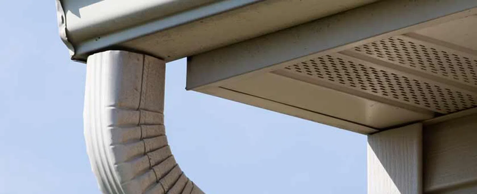 White gutter and downspout attached to the corner of a beige house under a blue sky.