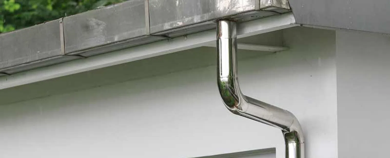 Modern house with shiny stainless steel rain gutter and downspout system on white facade under green trees.