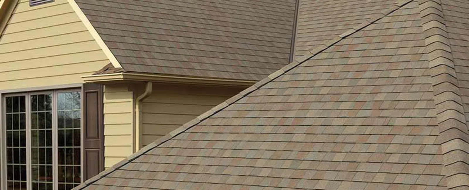 Close-up of residential house roof with brown shingles and stone chimney under a partly cloudy sky.