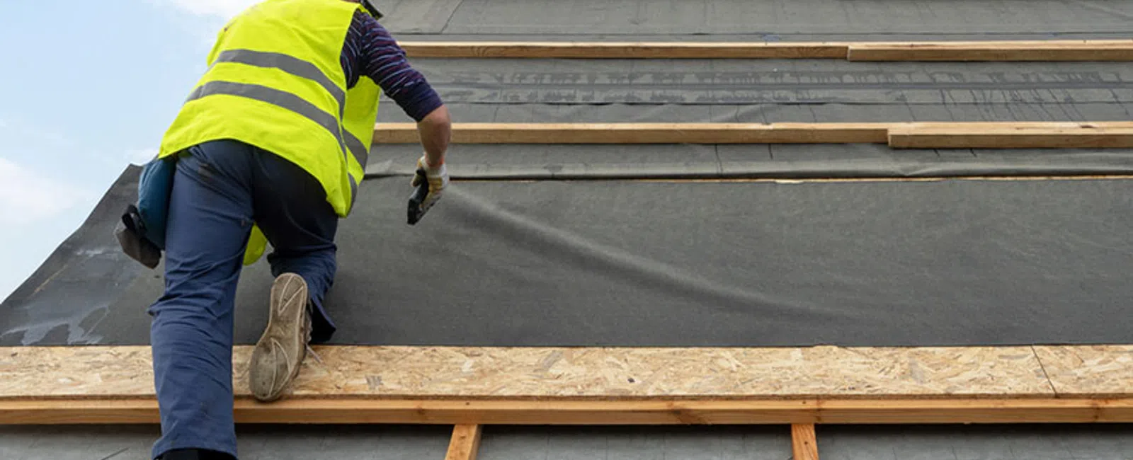 Construction worker in a high-visibility vest installing roofing underlayment on a wooden frame roof structure.