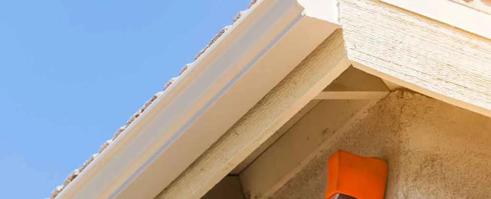 Close-up of roof corner with beige siding, white gutter, and orange ladder stoppers under clear blue sky.