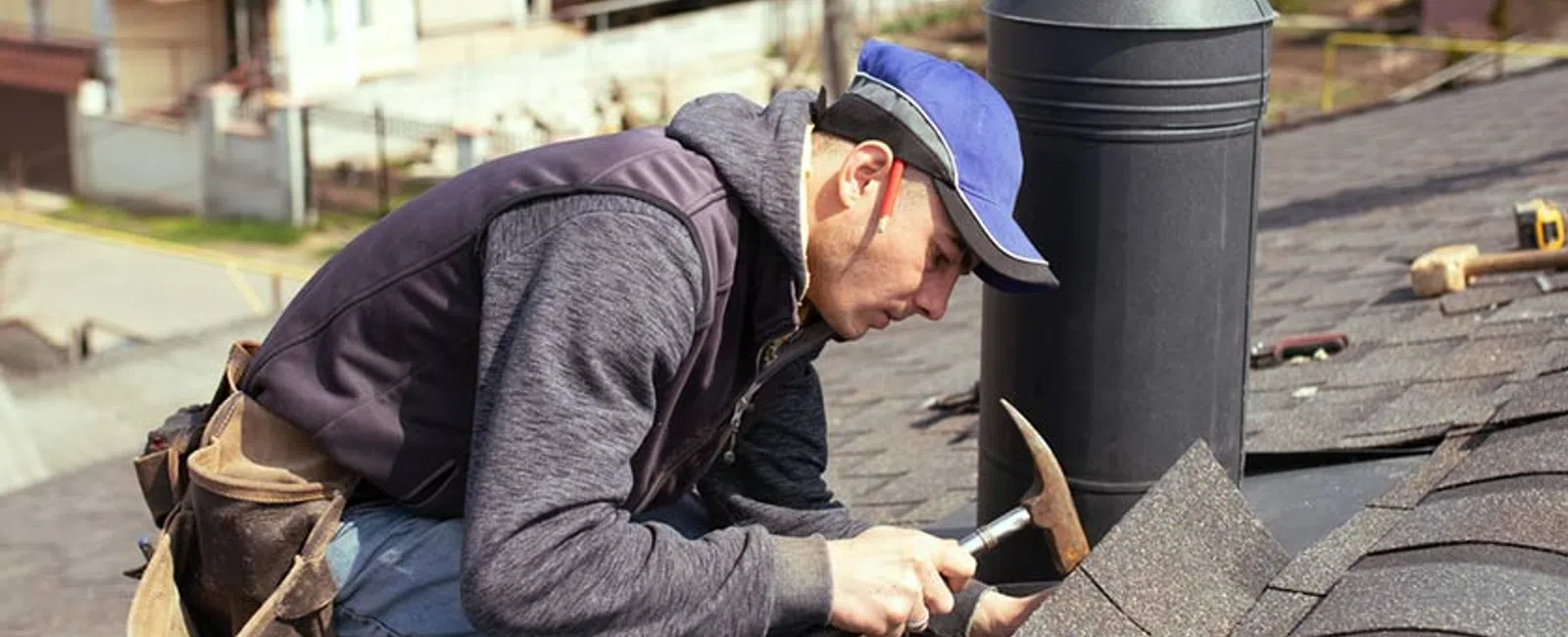 Roofer in blue cap installing shingles on rooftop using hammer near chimney pipe on a sunny day