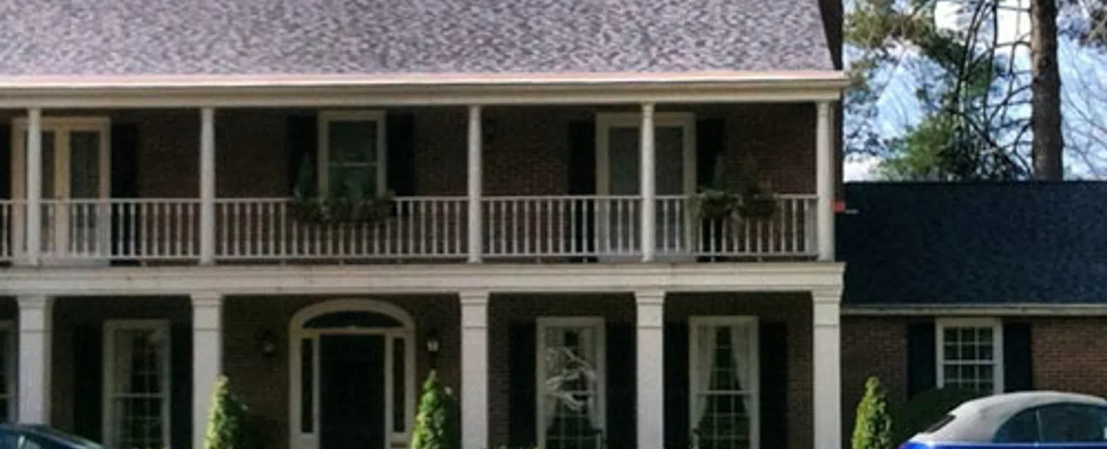 Two-story brick house with white columns, black shutters, balcony, and cars parked in front on a sunny day