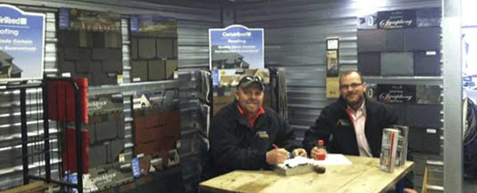Two men sitting at a table in a roofing materials showroom with shingles displayed on walls