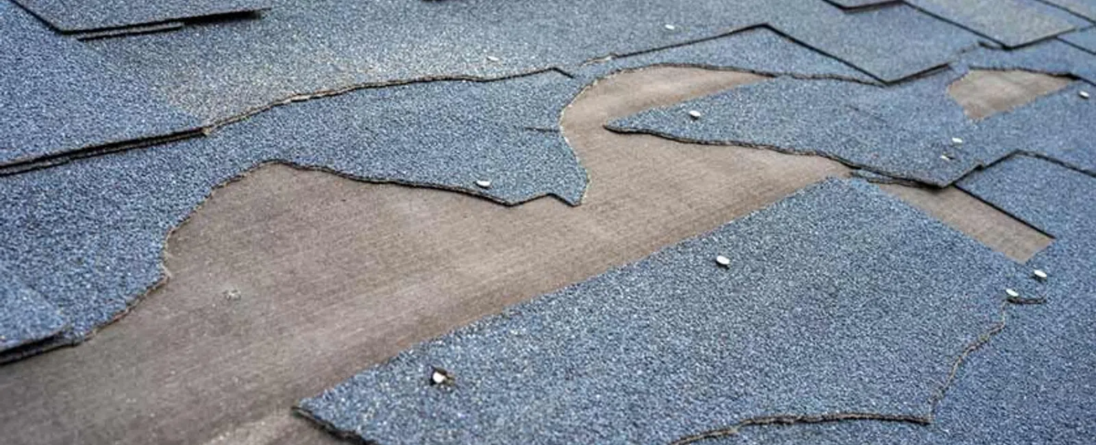 Damaged gray asphalt roof shingles peeling and missing, exposing the wooden roof deck underneath.