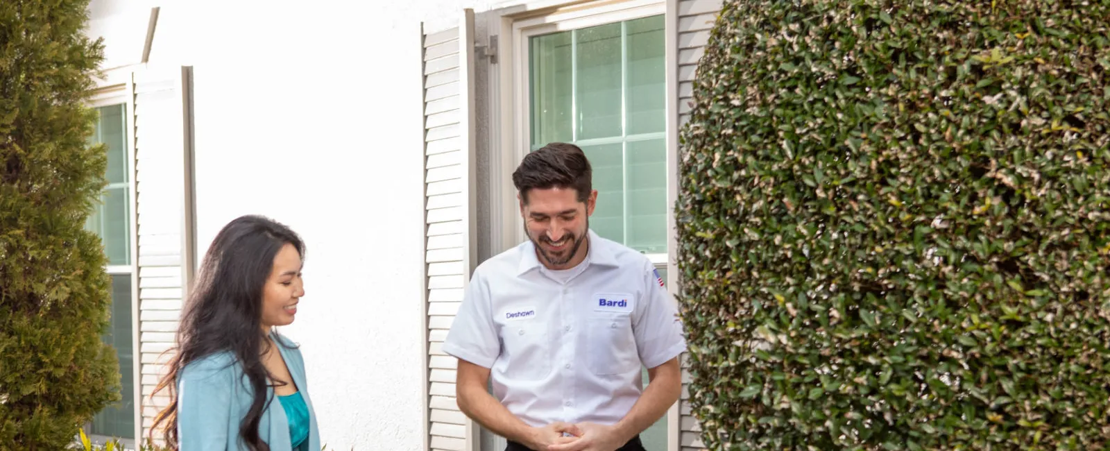 Technician explaining HVAC unit to woman outside house near trimmed tree and bushes on sunny day